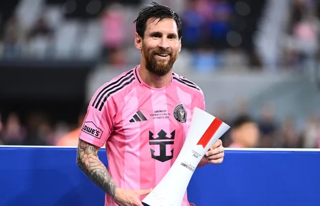 Professional soccer player with a pink jersey and beard, smiling while holding a rolled trophy or certificate at a stadium.
