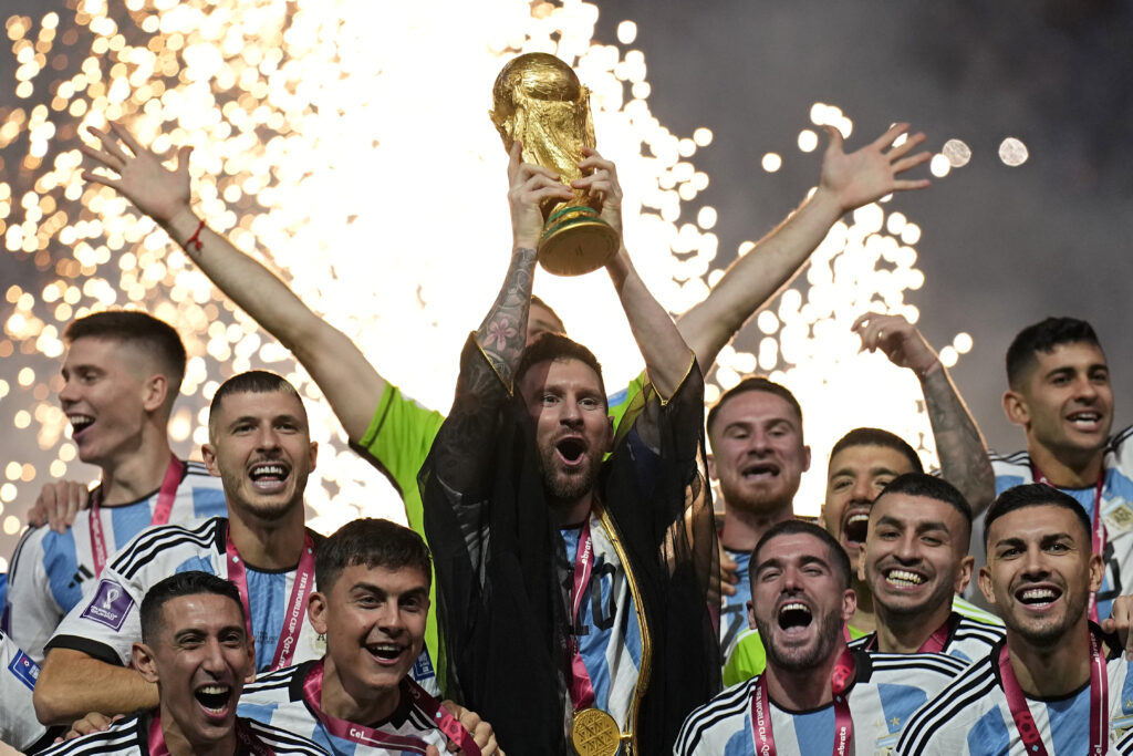 Soccer team lifts a gold trophy in celebration as bright fireworks explode in the background, players cheering and raising the trophy.