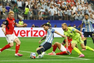 Soccer match action: players contest the ball near the goal line as a neon-yellow goalkeeper lunges to reach it. Crowd in the stands watches.