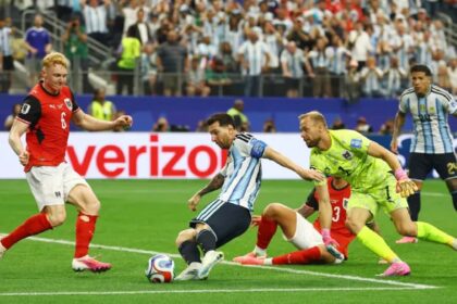 Soccer match action: players contest the ball near the goal line as a neon-yellow goalkeeper lunges to reach it. Crowd in the stands watches.
