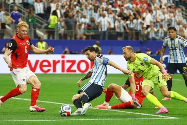 Soccer match action: players contest the ball near the goal line as a neon-yellow goalkeeper lunges to reach it. Crowd in the stands watches.