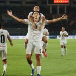 Soccer players in white celebrate on the field, one teammate carried on another's shoulders with arms outstretched behind them.