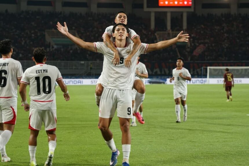 Soccer players in white celebrate on the field, one teammate carried on another's shoulders with arms outstretched behind them.