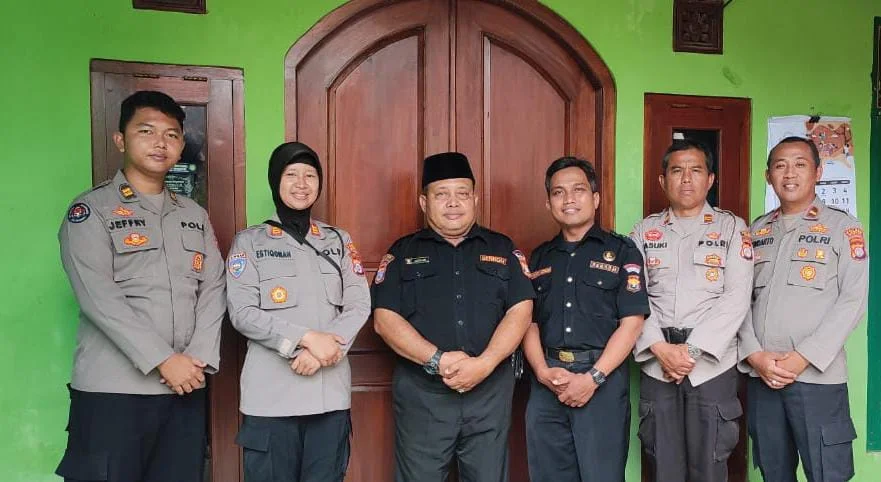 Six police officers pose for a group photo in front of a wooden door on a bright green wall; center figure wears black shirt and cap.