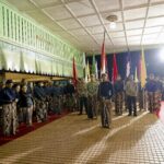 Group of people in batik attire standing in a line with colorful flags during a night ceremony under a green awning.