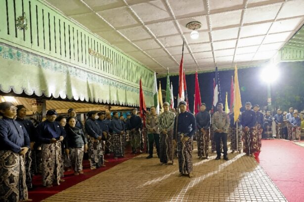 Group of people in batik attire standing in a line with colorful flags during a night ceremony under a green awning.