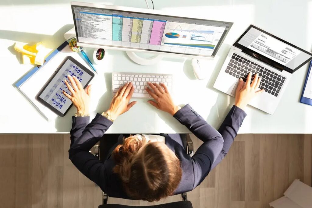 Top-down view of a person working at a glass desk with a laptop, a desktop monitor showing charts, a tablet, and a keyboard.