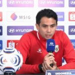 Indonesian footballer in a red jersey speaks into a blue microphone at a press conference, with a soccer ball on the table and sponsor backdrops behind him.