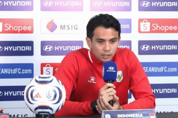 Indonesian footballer in a red jersey speaks into a blue microphone at a press conference, with a soccer ball on the table and sponsor backdrops behind him.