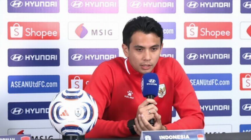 Indonesian footballer in a red jersey speaks into a blue microphone at a press conference, with a soccer ball on the table and sponsor backdrops behind him.