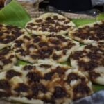Flatbreads topped with dark, seasoned meat arranged on banana leaves at a market stall
