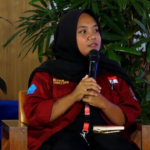Woman in a red jacket and black hijab seated, speaking into a handheld microphone at an event with green plants in the background