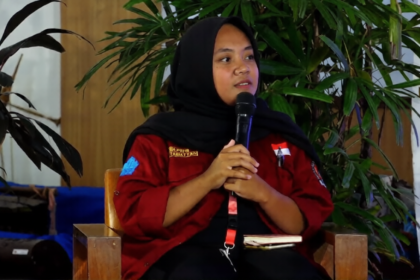 Woman in a red jacket and black hijab seated, speaking into a handheld microphone at an event with green plants in the background