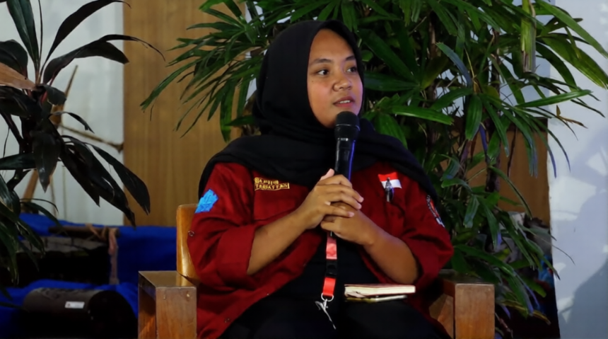 Woman in a red jacket and black hijab seated, speaking into a handheld microphone at an event with green plants in the background