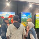 People in an art gallery viewing colorful landscape paintings on black walls.