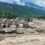 A village with colorful houses sits along a muddy riverbed after a flood, with ruined buildings in the foreground and green hills behind.