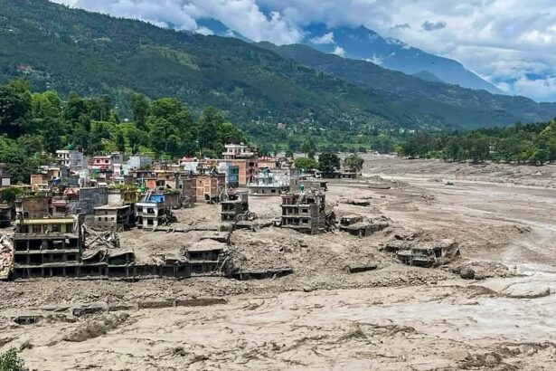 A village with colorful houses sits along a muddy riverbed after a flood, with ruined buildings in the foreground and green hills behind.