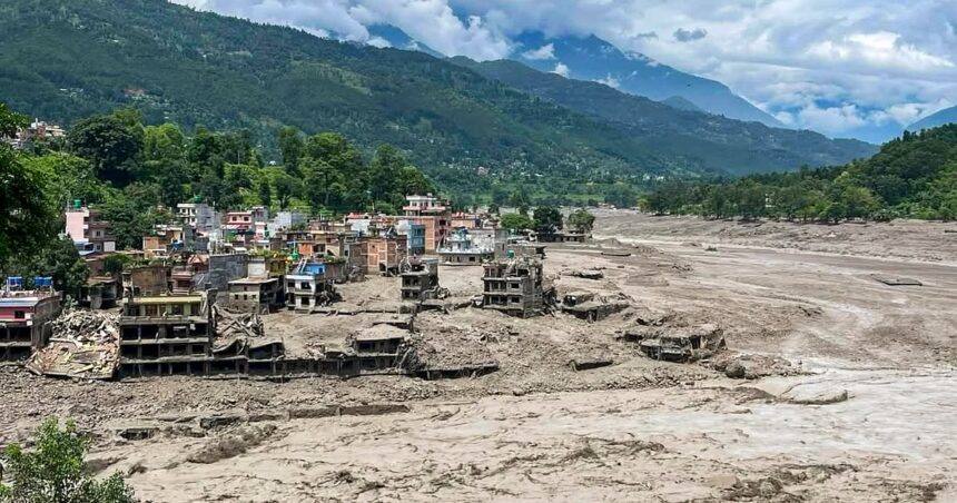 A village with colorful houses sits along a muddy riverbed after a flood, with ruined buildings in the foreground and green hills behind.