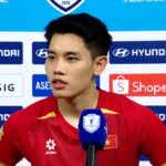 Young Asian football player in a red jersey giving a post-match interview in front of a sponsor backdrop.