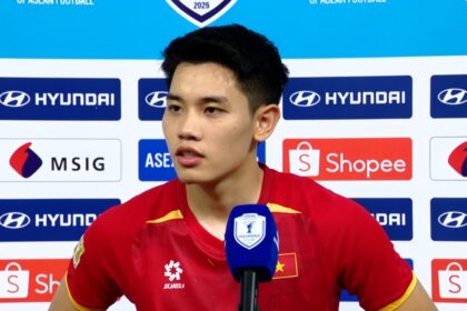 Young Asian football player in a red jersey giving a post-match interview in front of a sponsor backdrop.