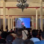 Crowd seated in a large hall watching a motorcycle race on a big screen, surrounded by cream pillars and a chandelier in the ceiling.