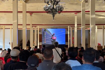 Crowd seated in a large hall watching a motorcycle race on a big screen, surrounded by cream pillars and a chandelier in the ceiling.