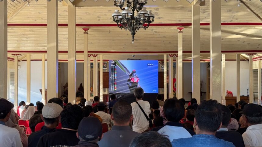 Crowd seated in a large hall watching a motorcycle race on a big screen, surrounded by cream pillars and a chandelier in the ceiling.