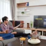 Couple relaxing on a gray sofa in a bright living room, watching TV with snacks and drinks nearby.