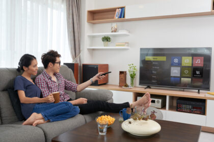 Couple relaxing on a gray sofa in a bright living room, watching TV with snacks and drinks nearby.