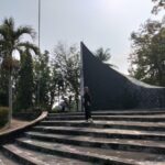 Person standing on wide concrete steps in front of a tall, curved dark monument in a park with palm trees and other greenery behind it.