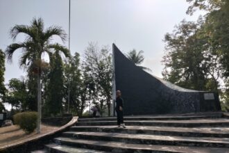 Person standing on wide concrete steps in front of a tall, curved dark monument in a park with palm trees and other greenery behind it.