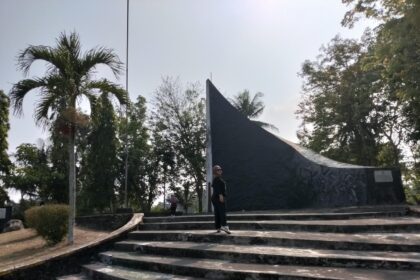 Person standing on wide concrete steps in front of a tall, curved dark monument in a park with palm trees and other greenery behind it.