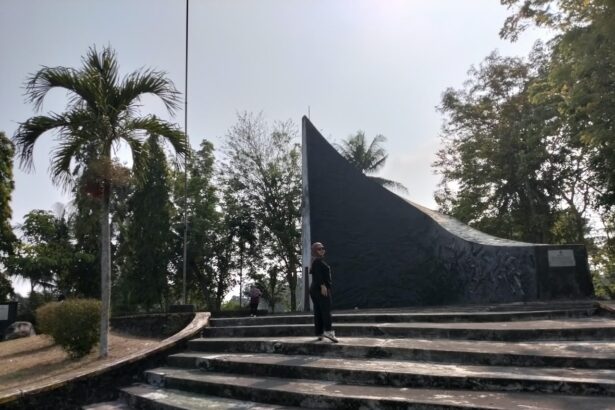 Person standing on wide concrete steps in front of a tall, curved dark monument in a park with palm trees and other greenery behind it.