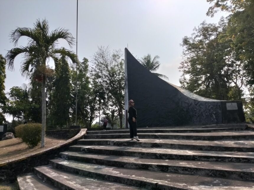 Person standing on wide concrete steps in front of a tall, curved dark monument in a park with palm trees and other greenery behind it.