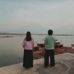 Two people stand on a concrete edge, facing a calm harbor with docked wooden boats seen in the water.