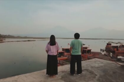 Two people stand on a concrete edge, facing a calm harbor with docked wooden boats seen in the water.