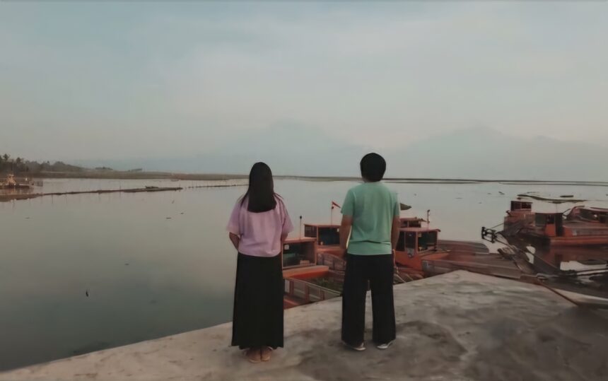 Two people stand on a concrete edge, facing a calm harbor with docked wooden boats seen in the water.