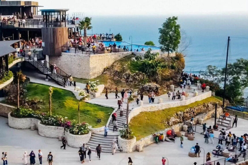 Terraced seaside resort with people on patios overlooking the ocean, a pool, and stone paths.