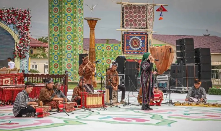 Group of musicians in traditional attire performing gamelan on an outdoor stage with colorful fabric panels behind them