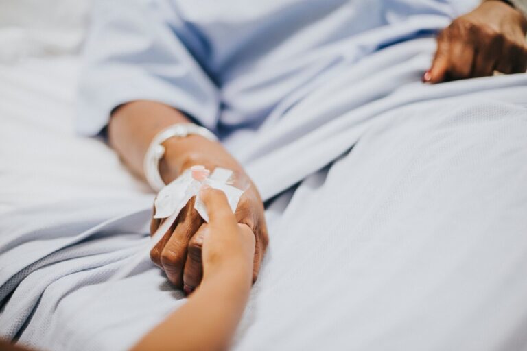 Hospital patient reaches out to hold a caregiver's hand; IV line and medical bracelet visible on the wrist in a hospital bed.