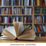 Open book spread fanned on a wooden table with shelves of books in a library in the background.