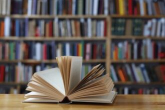 Open book spread fanned on a wooden table with shelves of books in a library in the background.