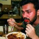 Man with a beard and red sauce on his lips eats with a spoon at a booth in a casual restaurant, surrounded by other diners at wooden tables.