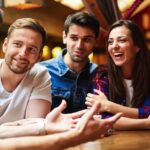 A group of six friends sits around a wooden table, laughing and chatting over coffee in a cozy cafe.