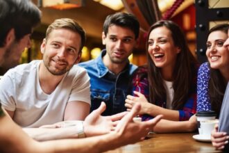 A group of six friends sits around a wooden table, laughing and chatting over coffee in a cozy cafe.