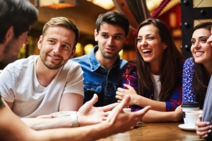 A group of six friends sits around a wooden table, laughing and chatting over coffee in a cozy cafe.