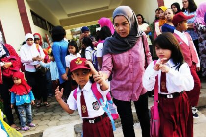 Group of children in uniforms with backpacks outside a school, adults nearby awaiting drop-off.
