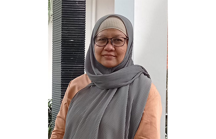 Woman wearing a gray hijab and glasses, standing beside a white column outdoors and smiling at the camera.