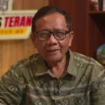Man with glasses in a green patterned shirt speaks on camera in a studio, shelves visible in the background.