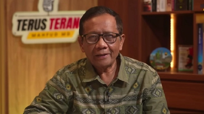 Man with glasses in a green patterned shirt speaks on camera in a studio, shelves visible in the background.